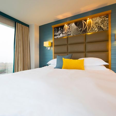 Voco St. David'S Cardiff By Ihg Hotel Buitenkant foto The photo shows a modern hotel room featuring a large bed with a neatly made white duvet. The headboard is upholstered in a soft gray material, complemented by decorative lighting on either side. Above the headboard, there is a stylish wall art displ