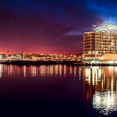 Voco St. David'S Cardiff By Ihg Hotel Buitenkant foto The photo depicts a modern building, likely a hotel or office complex, situated by a waterfront. It features a distinctive architectural design with a curved roof and multiple levels of glass windows. The scene is captured during twilight, creating a