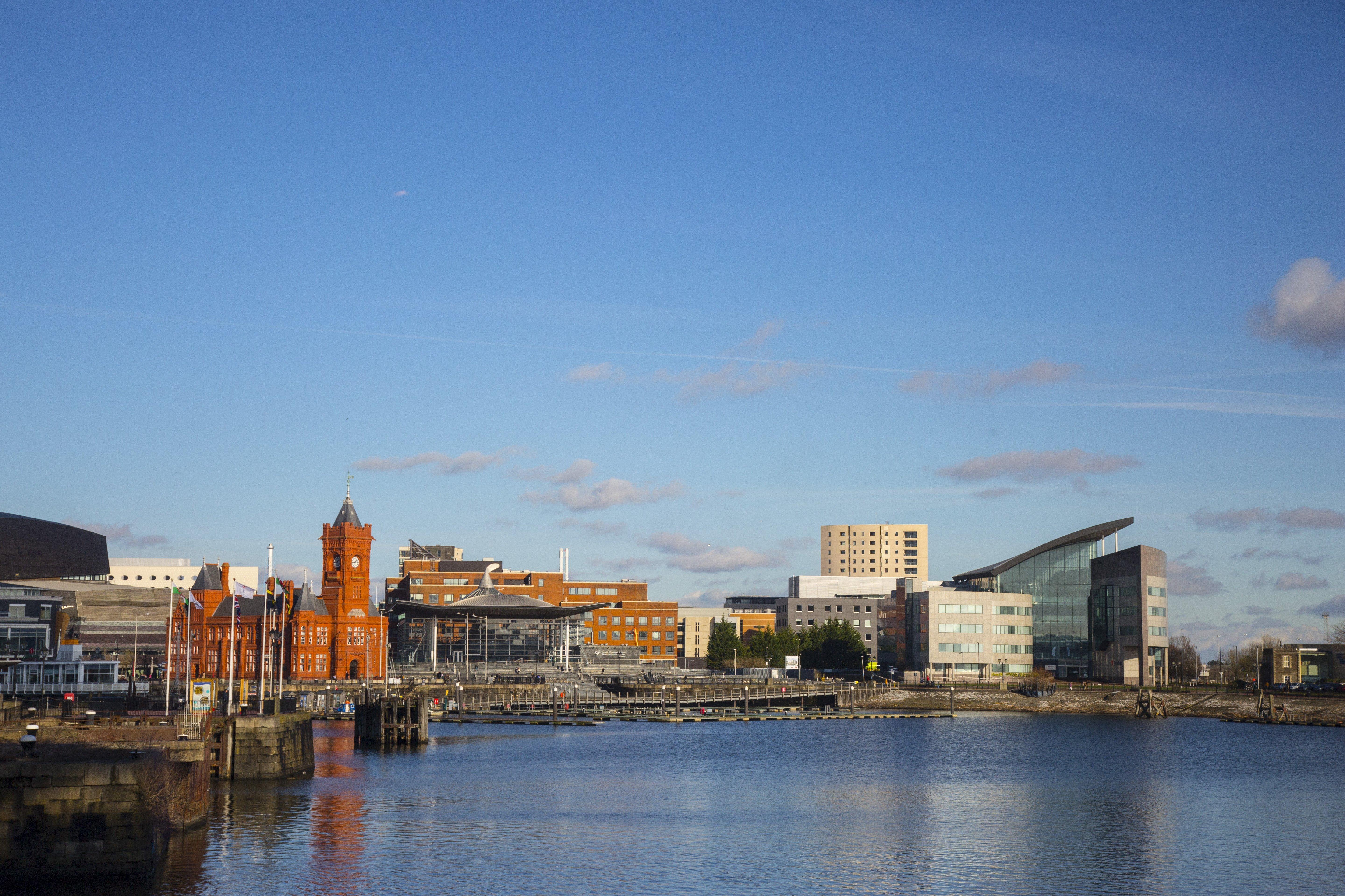 Voco St. David'S Cardiff By Ihg Hotel Buitenkant foto The photo depicts a picturesque waterfront scene featuring a mix of architectural styles. On the left side, there is a prominent red brick building with a tower, which is likely a historical structure. To the right, modern buildings with sleek design
