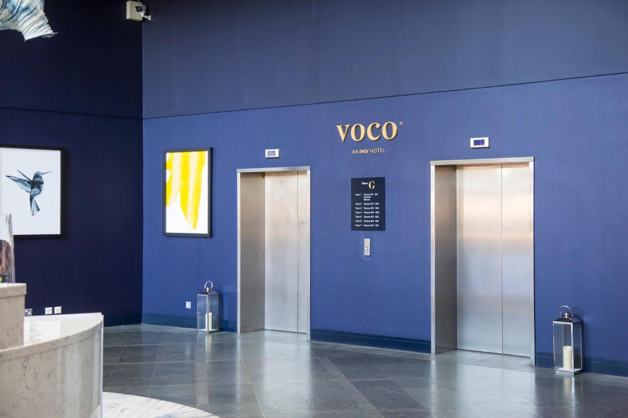 Voco St. David'S Cardiff By Ihg Hotel Buitenkant foto The image shows an interior space, likely a lobby of a building or hotel branded as "Voco." The walls are painted a deep blue color. There are two elevators with metallic doors, positioned side by side. Above the elevators, there is a sign that displ